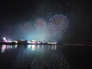 Un feu d'artifice éclate dans le ciel nuit noir sous les bâtiments du centre-ville de Martigues, et l'eau du canal en premier plan. Les reflets des couleurs roses et vertes se reflètent dans l'eau