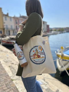 Une jeune fille de profil est debout sur le site du Miroir aux Oiseaux à Martigues. Elle porte un sac en tissu et une gourde sur lesquels figurent un nouveau logo dessiné