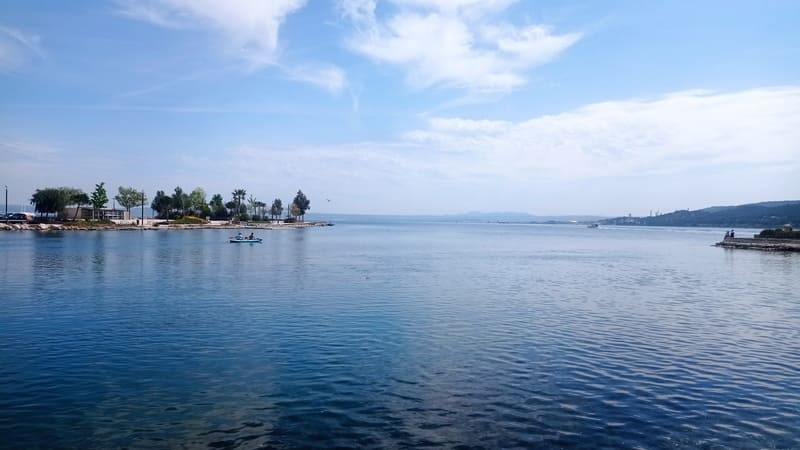 Etang de Berre et sa vaste étendu d'eau avec des arbres sur ses abords et un kayak qui glisse sur l'eau. Les nuages dans le ciel amène un aspect paisible à la photo