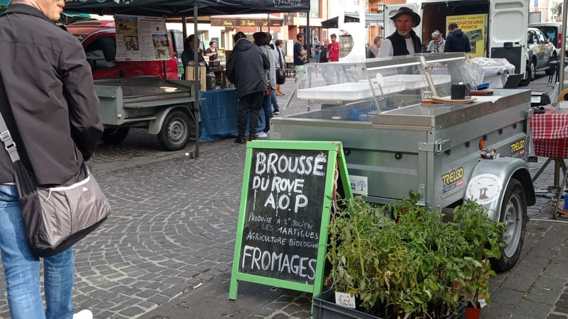 Au premier plan, un visiteur du marché des producteurs locaux de Martigues. Un panneau annonce la vente de brousse du Rove AOP. En fond, des stands et des visiteurs dans les allées sont visibles