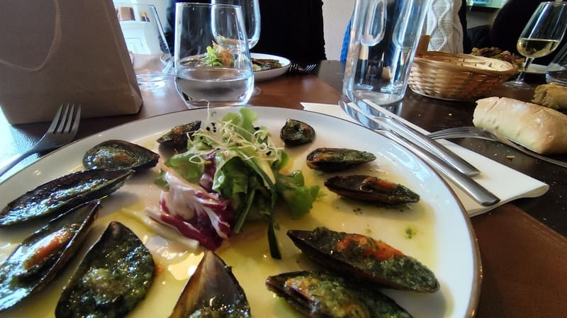 Assiette de moules gratinées en persillade avec sa sauce marinière. Les moules sont ouvertes et l'assiette ronde posée sur une table en bois. Des couleurs sont disposés à côté des verres
