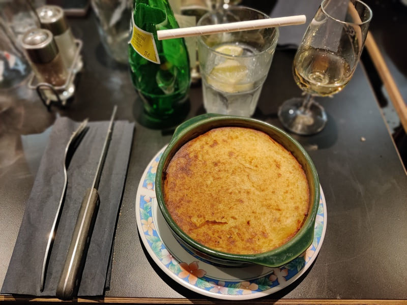 Une assiette ronde est agrémenté d'un bol en terre cuite. Le bol est rempli de hachis parmentier. Des couverts, des verres et des bouteilles sont disposés sur la table noire
