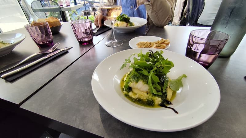 Un assiette pour trois couverts avec des verres et des assiettes blanches. Les assiettes contiennent du poisson blanc avec de la salade, la lumière du jour éclaire la tablée