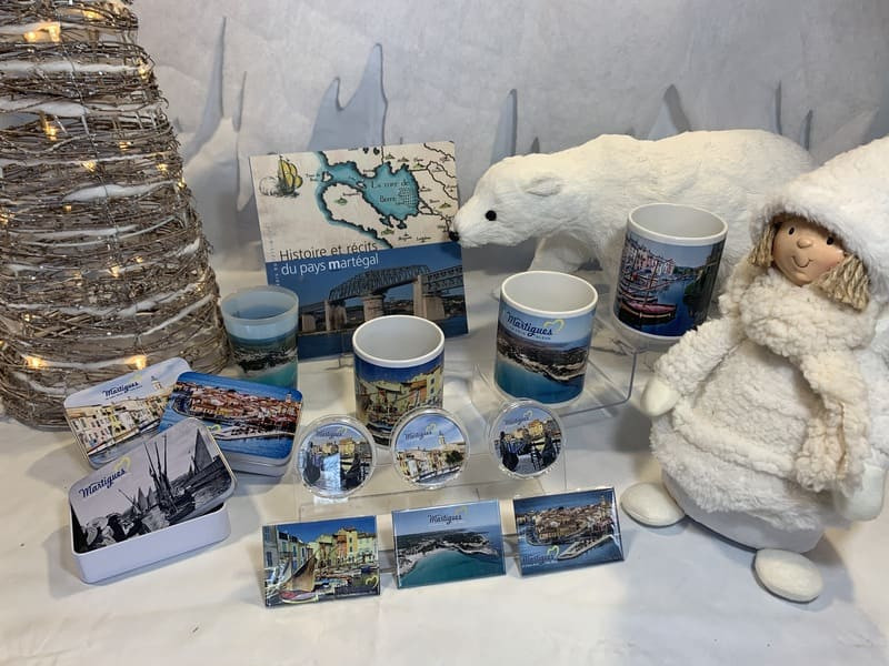 Présentation des produits de la gamme Martigues avec boîtes, mugs, magnets, livres et gobelets. Le tout entouré de décorations de Noël blanches.