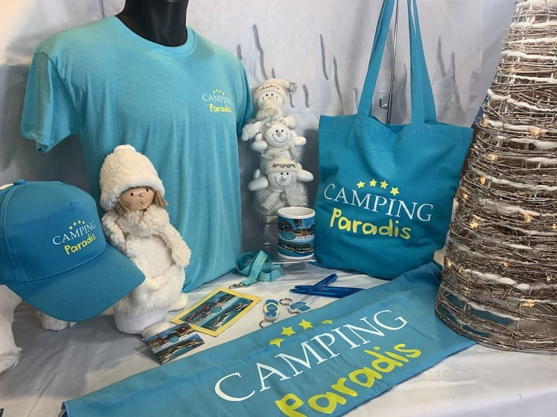 Jolie présentation des produits de la gamme Camping Paradis. Bleu turquoise et jaune qui rappelle les vacances. Tote bag, serviette, tee-shirt, casquette, mug, magnet et tour de cou sont en vente à l'Office de Tourisme de Martigues
