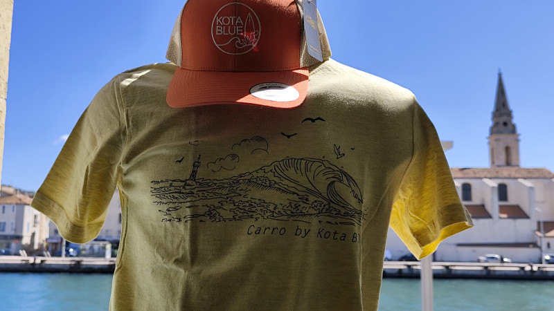 Tee-shirt jaune de la marque Kota Blue avec dessin d'une vague et du phare de La Couronne. Casquette orange et blanche posée sur un mannequin avec en fond un paysage de Martigues