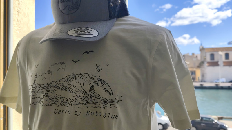 Tee-shirt blanc avec les symboles de la Côte Bleue : vague, surf, mouettes et phare de La Couronne. Une casquette bleue et grise avec le logo de la marque est posée sur le tee-shirt