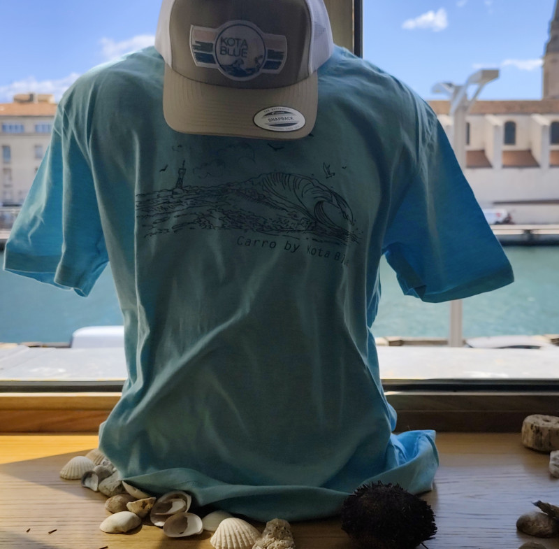 Tee-shirt bleu avec motif d'une vague et des mouettes autour d'un phare de côte méditerranéenne. Casquette beige et blanche avec logo de Kota Blue en couleur