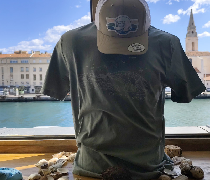 Tee-shirt gris avec dessin en blanc d'une vague et de mouettes volant dans le ciel. Une casquette beige et blanche avec le logo de la marque. Le buste du mannequin est posé au milieu de coquillages