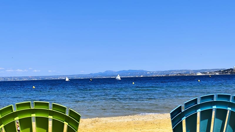 Deux transats aux couleurs vives sont disposés de dos face à la mer sur la plage de Ferrières à Martigues. Le ciel est bleu sans nuage, les bateaux voguent sur l'étang de Berre