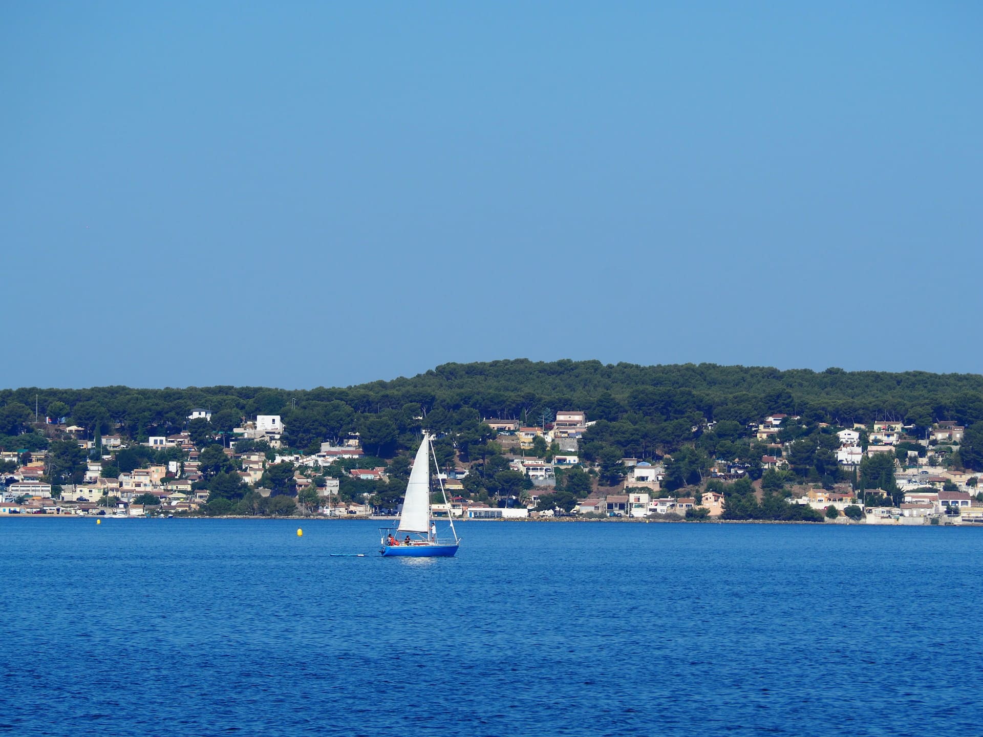 The sea of the Etang Bleu with a sailboat and its white sail in its center. In the background, the lagoons and green hills with the residential houses around the Etang de Berre on  - © Otmartigues / EstelleB