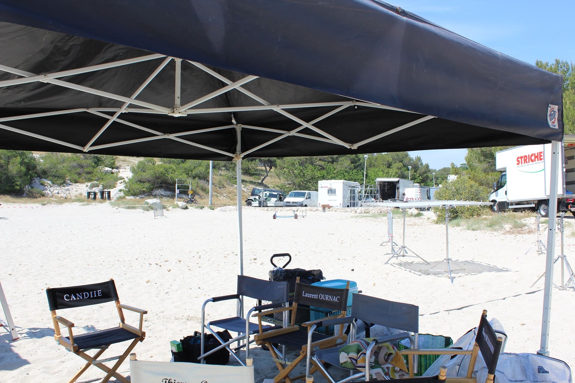 Fauteuils de tournage sous un barnum lors du tournage de la série télévisée Camping Paradis sur la plage de Sainte-Croix. Au loin, des camions de production sont installés - © Otmartigues / ElodieM