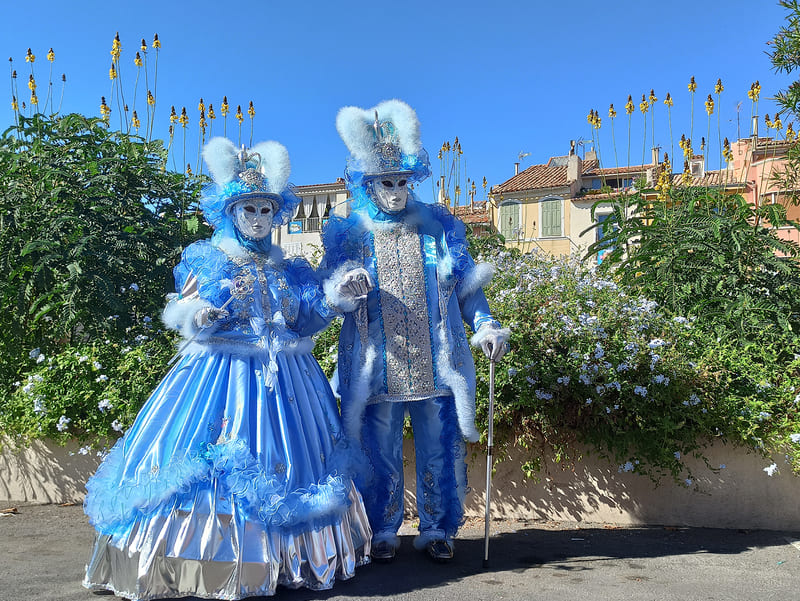 Pareja de figuras enmascaradas venecianas de colores a juego. La dama lleva un amplio vestido con volantes, sombrero y abanico. El caballero lleva un traje con bastón y sombrero. A - © Otmartigues / KarimK