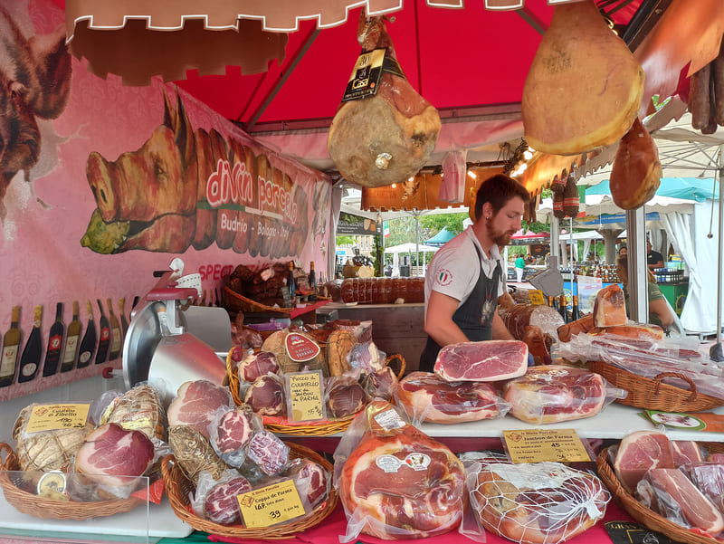 Puesto de charcutería italiana en el mercado de Les Italiennes, celebrado en septiembre en Martigues. Se presentan embutidos en cestas y jamones enteros cuelgan del techo. - © Otmartigues / KarimK
