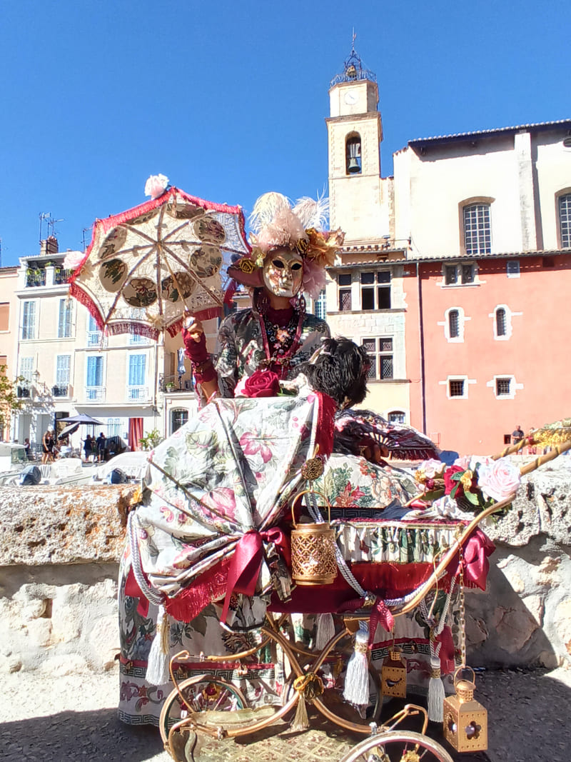 Personaje veneciano enmascarado con traje tradicional de muñeca, bolsa de equipaje, sombrilla y cochecito de bebé antiguo desfilando por las calles de Martigues. - © Otmartigues / KarimK