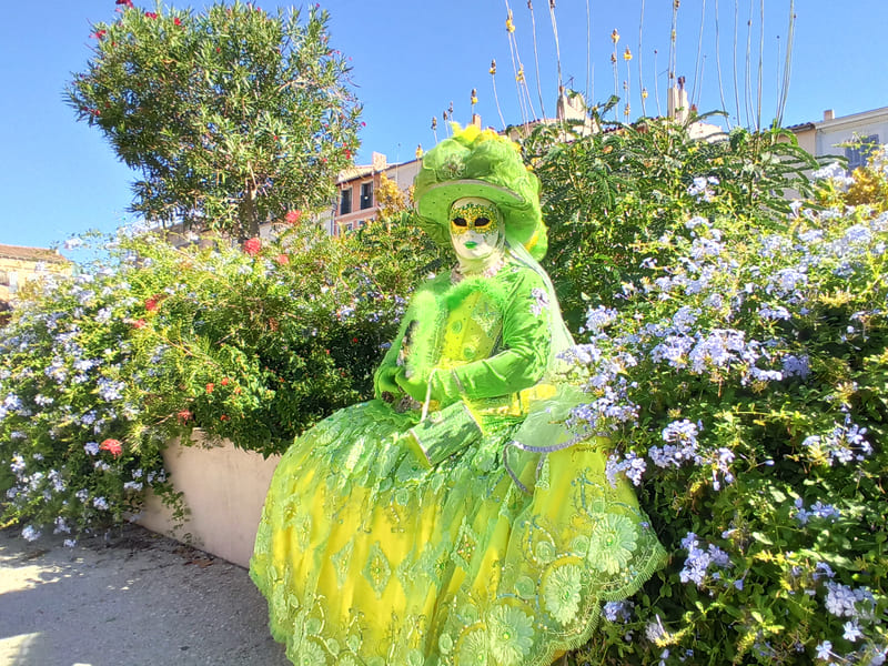Lleva un llamativo vestido verde con volantes y el rostro completamente enmascarado, con un gran sombrero armonioso, sentado sobre un muro bajo en flor. - © Otmartigues / KarimK
