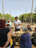 Les participants à l'atelier cuisine sont assis sur des tables de pique-nique et entourent le chef Julien qui dévisse une bouteille d'huile d'olive pour préparer des sandwichs. Des pains bagnats sont posés sur un plateau