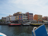 Photo prise depuis un bateau sur les canaux de Martigues. On doit une aile bleue du bateau au premier plan. Au second plan, les maisons aux façades colorées et les barques traditionnelles des rameurs vénitiens