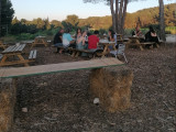Gente sentada de espaldas en mesas de picnic en el centro de la granja. El suelo está cubierto de hojas, rodeado de árboles. El cielo es azul y brillante.