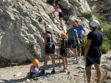 Le groupe de personne s'initie à l'escalade dans les calanques de la Côte Bleue avec BNN et l'Office de Tourisme de Martigues. L'activité est ouverte aux adultes et aux enfants. Une personne commence à gravir la roche.