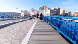 Des jeunes filles de dos marchent et traversent un pont bleu dont le sol est en pavé. Au bout, des bâtiments aux façades colorées. De part et d'autres du pont, l'eau.