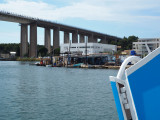 La porte d'embarquement du bateau est bleue au premier plan. On voit le viaduc autoroutier. Dessous, des cabanes de pêcheurs en référence au Calen de Martigues.
