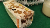 Un nougat blanc dans son emballage posé sur fond vert. Des gros morceaux d'amandes sont visibles, ce qui confère au produit son naturel et donne envie de croquer dedans