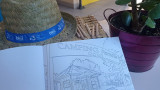 Carnet de coloriage ouvert montrant un dessin de Camping Paradis, entouré d'un chapeau de paille et d'une plante