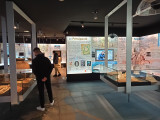 Hall principal de la galerie de l'Histoire où deux jeunes hommes regardent les vitrines. Des grands panneaux informatifs avec des cartes sont placardés aux murs.