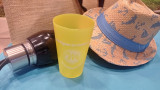 Gelber Becher aus der Gamme Soleil mit dem Logo des Fremdenverkehrsamts von Martigues, auf einem blauen Strandtuch platziert, zusammen mit einem Strohhut, einer Strandtasche und einer Wasserflasche