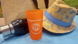 Orange cup from the Soleil range featuring the logo of the Martigues Tourist Office, placed on a blue beach towel alongside a straw hat, a beach bag, and a water bottle