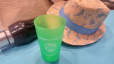 Green cup from the Soleil range featuring the logo of the Martigues Tourist Office, placed on a blue beach towel alongside a straw hat, a beach bag, and a water bottle
