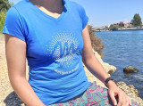 Tee-shirt femme bleu de la Gamme Soleil, orné du logo blanc de l'Office de Tourisme au centre, porté par un modèle féminin avec vue sur le canal en arrière-plan