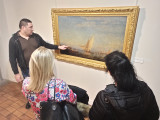 Deux femmes positionnées de dos écoutent les explications d'un homme guide. Il présente un tableau célèbre de Félix Ziem représentant un voilier sur l'étang de Berre.