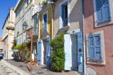 Zoom sur les façades colorées aux couleurs pâles du Miroir aux Oiseaux. Les volets et portes des maisons sont bleus. Un lampadaire à la forme typique de Provence est au premier plan. Sur le côté, on devine un début de la façade de l'église.