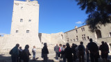 Le groupe est dans la cour du Fort. Une tour se trouve à gauche. Des escaliers et un bâtiment à droite. Un arbre au premier plan. Un jeu de lumière avec soleil et ombre.