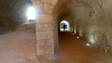 Dans les couloirs du Fort, deux passages souterrains avec de la lumière au bout. Des personnes sont engagés dans le tunnel de droite. Le sol est en terre, les tunnels en pierre de couleur ocre.