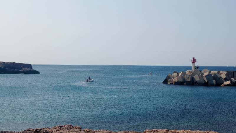 Vu de l'horizon depuis le village de Carro. Un phare rouge et blanc est présent sur le digue. Un bateau s'élance vers la mer méditerranée. Des rochers sont présents à nos pieds et sur la droite.
