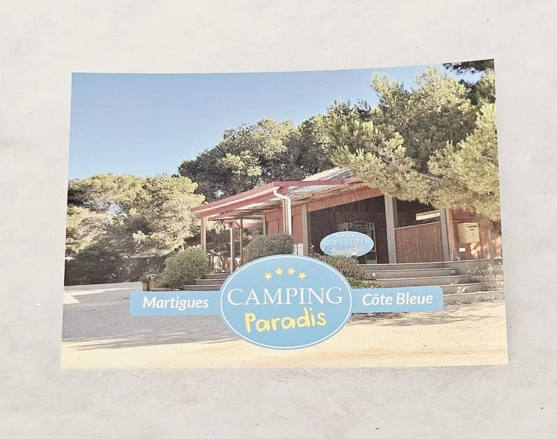 Postal horizontal con un primer plano de la entrada del supermercado, un conjunto de la serie Camping Paradis. El logotipo de la serie se encuentra en la mitad inferior de la postal. - © Otmartigues / MarianneB Postal que representa el icónico set de la serie Camping Paradis: el supermercado. Las postales se pueden comprar durante las visitas a los lugares de rodaje de la serie o en la Oficina de Turismo de