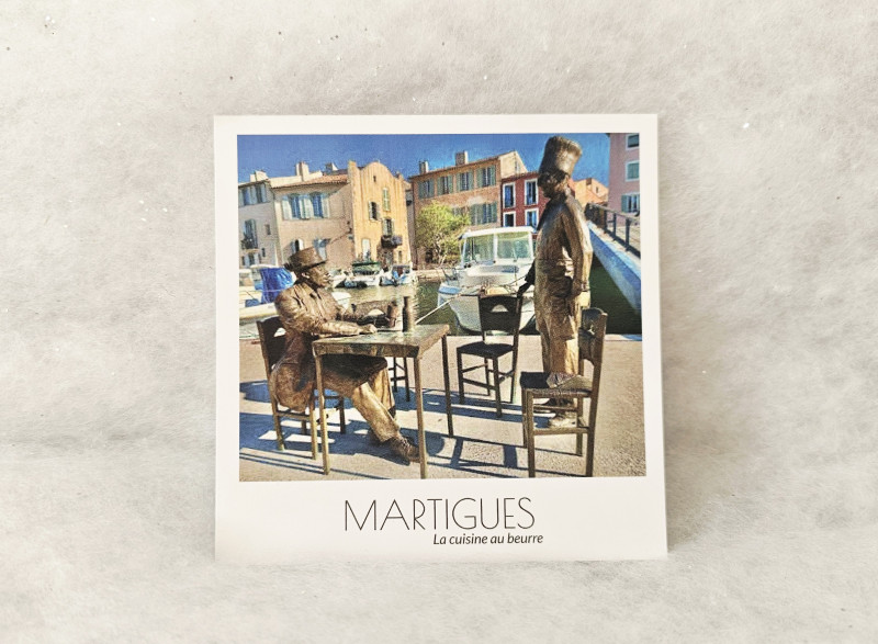Postal cuadrada que representa la escultura de Bourvil y Fernandel sentados a una mesa, un homenaje a la película La Cuisine au Beurre, en el distrito de la Île de Martigues. - © Otmartigues / MarianneB Postal estilo Polaroid de la estatua de bronce de Bourvil y Fernandel en Martigues, distrito de la Île. Una referencia a la película de culto La Cuisine au Beurre (La Cocina de la Mantequilla), film