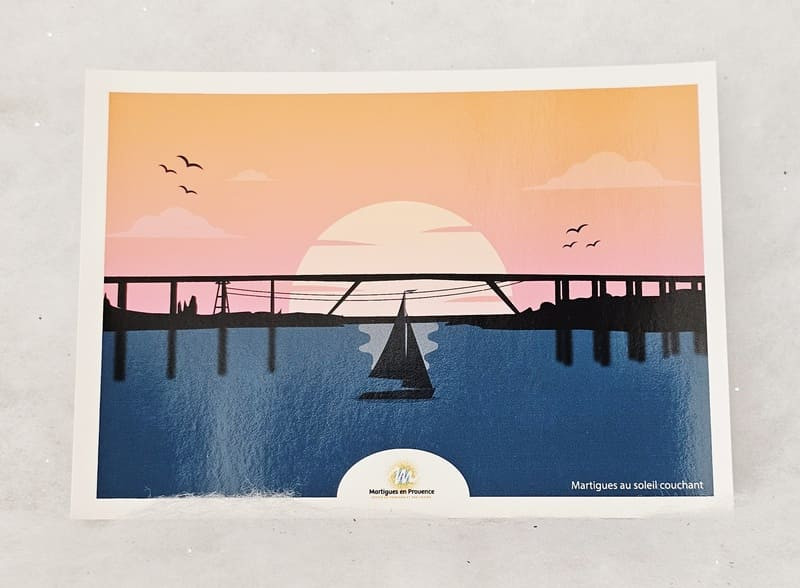 Illustrated postcard depicting a sailboat on the Black Sea, with the motorway viaduct in the background and a glowing red sky illuminated by a setting sun. - © Otmartigues / MarianneB Postcard featuring a drawing of the viaduct and sailboats crossing the Martigues canal. A souvenir available for purchase at the Martigues Tourist Office shop.