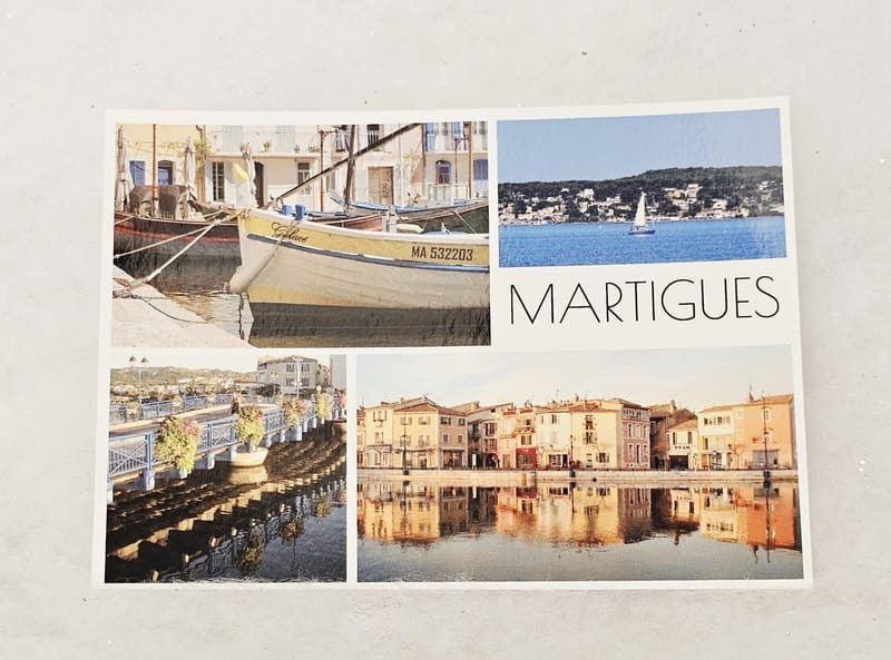 Postal con varias fotos que representan imágenes icónicas del centro de Martigues: un puente, barcos, casas de colores y un velero en el Étang de Berre. El nombre de Martigues aparece en la esquina su - © Otmartigues / MarianneB Una postal condensada del centro de Martigues, que incluye lugares patrimoniales, canales y el Étang de Berre. Puede adquirir una postal de recuerdo de Martigues en la Oficina de Turismo.