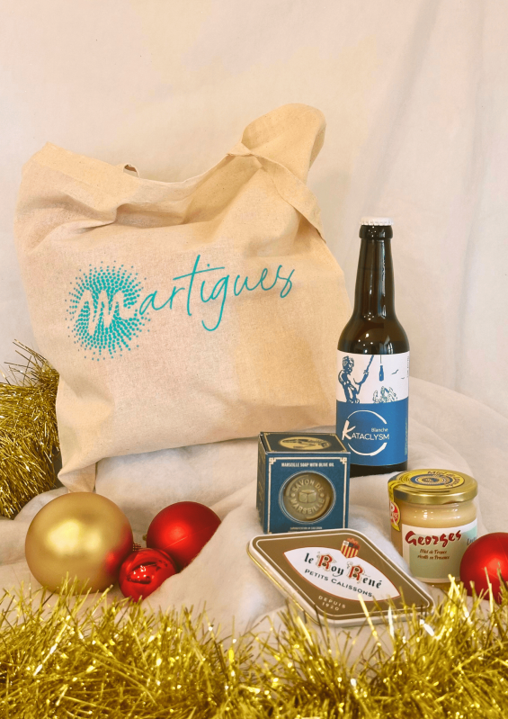 Coffret de Noël en vente à la boutique de l'Office de Tourisme de Martigues avec bière artisanale  - © Otmartigues / PaolaM Un coffret de Noël en vente à l'Office de Tourisme de Martigues comprenant un tote bag à l'effigie de Martigues, une bière artisanale Kataclysm, un savon Marius Fabre, une boîte de calissons Roy René, un pot de miel 125g Les Ruchers de Georges