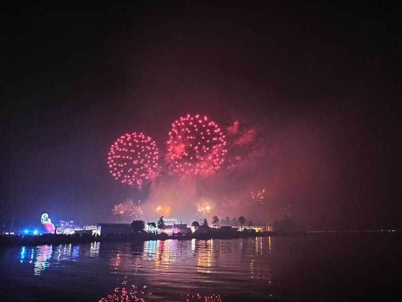 The red fireworks burst into the dark night sky. The Martigues church steeple is recognizable in the background. The water is calm, with reflections in it.