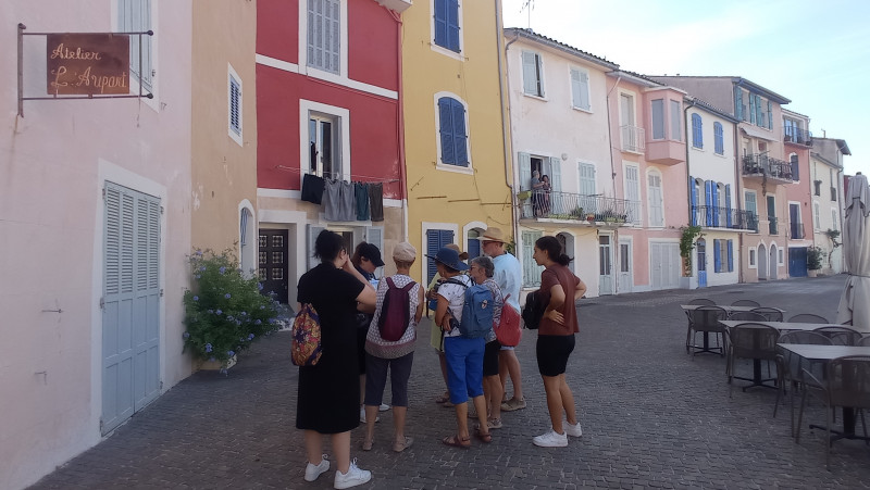 Un groupe de personnes est rassemblé sur une place dont les façades des maisons sont colorées. Des tables et chaises sont à droite de l'image. Une enseigne rouillée d'un atelier est attaché contre le mur à gauche.