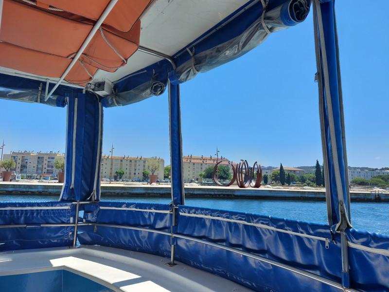 La photo est prise de l'intérieur d'un bateau au bâches transparentes. On voit la mer bleue et des sculptures en fer forgés représentant des cercles posées sur un quai en forme de proue d'un bateau.