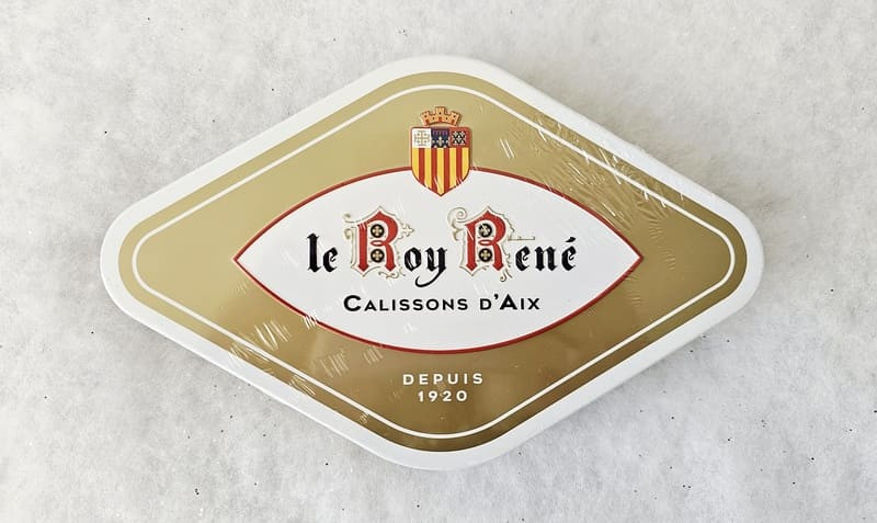 Esta caja de calissons dorada y blanca con forma de diamante evoca la clásica silueta del calisson. El logotipo de Roy René está impreso en letras en el centro. - © Otmartigues / MarianneB Caja de 18 calissons Roy René, fabricados en Aix-en-Provence, cerca de Martigues. Los productos Roy René están disponibles en la tienda de la Oficina de Turismo de Martigues.