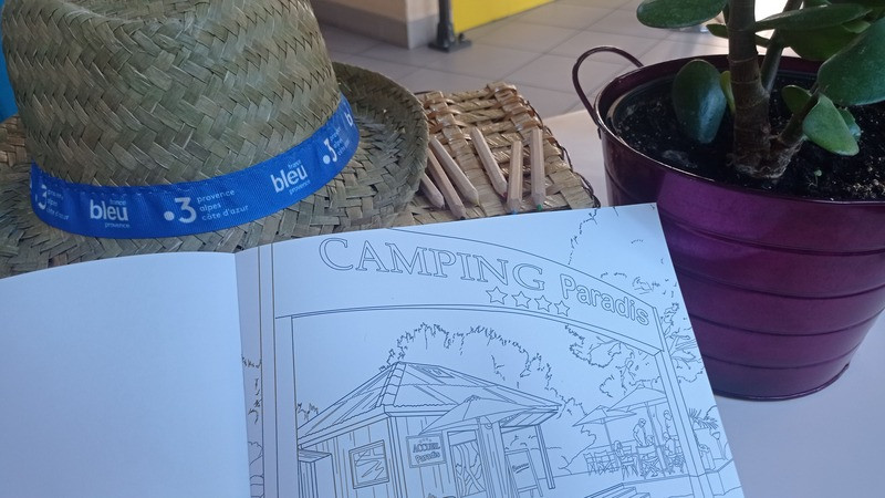 Carnet de coloriage ouvert montrant un dessin de Camping Paradis, entouré d'un chapeau de paille et d'une plante