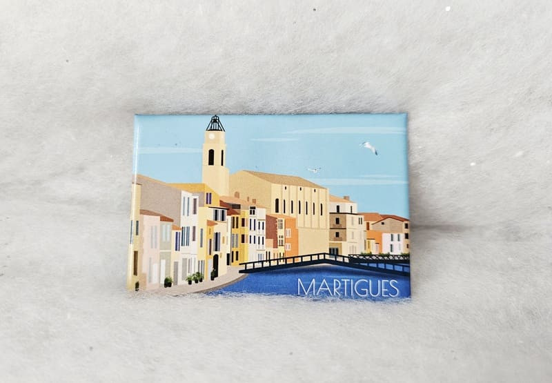 Ein Magnet mit einer Zeichnung des Kirchturms und der farbenfrohen Hausfassaden im Stadtzentrum. Verschiedene Magnete sind im Fremdenverkehrsamt von Martigues erhältlich.