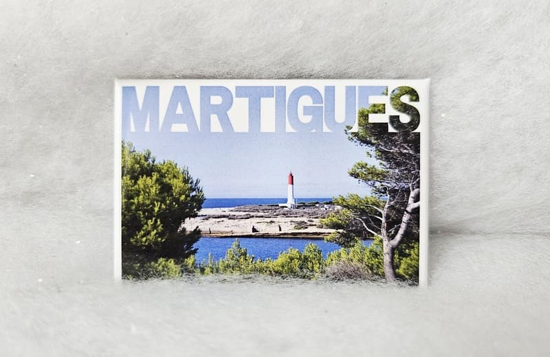 Imán con el texto Martigues incrustado en el cielo azul de la foto que representa la Costa Azul y el faro de La Couronne. Roca caliza, vistas al mar y pinos mediterráneos, típicos del sur de Francia - © Otmartigues / MarianneB Imán con la foto del faro de La Couronne en la Costa Azul de Martigues. Regalo de recuerdo disponible en la Oficina de Turismo de Martigues.